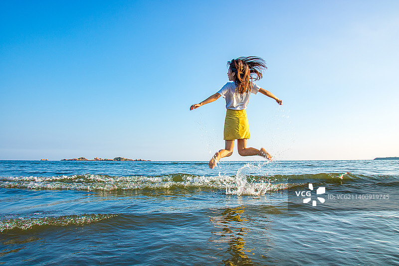 蓝天下一个在海边跳跃的长发女生图片素材