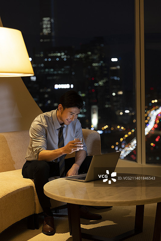 夜晚商务男子在酒店房间使用笔记本电脑视频，喝威士忌干杯图片素材