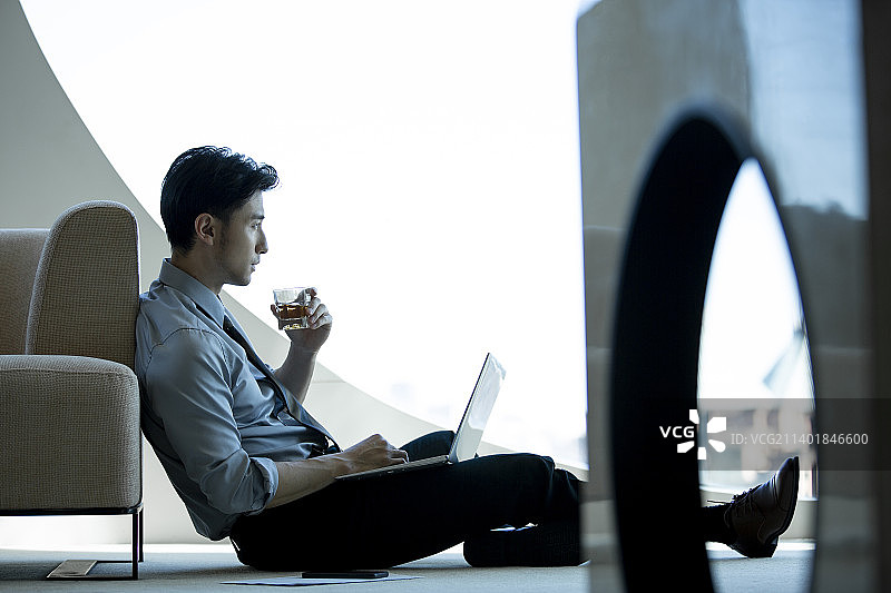 商务男子坐在酒店房间地上喝威士忌，使用笔记本电脑工作图片素材