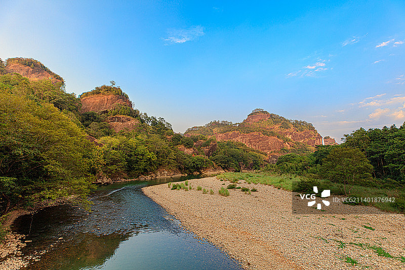 蓝天夕阳映照下的武夷山群峰，丹霞地貌在傍晚阳光下发出红光，九曲溪两侧绿树成荫，旱季溪水浅露出石子河滩图片素材