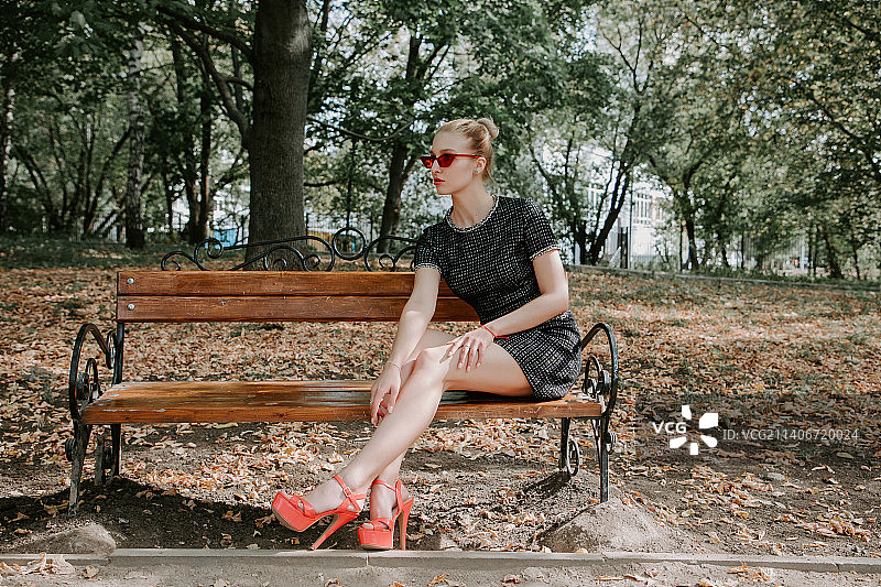 戴着红色太阳镜和黑色短裙坐在公园长椅上的漂亮金发女人图片素材