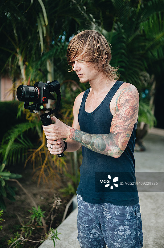 在泰国帕岸岛，手持稳定器的年轻男子站在树木周围的人行道上图片素材