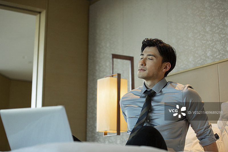 商务男子坐在酒店房间床上工作后闭着眼睛休息图片素材