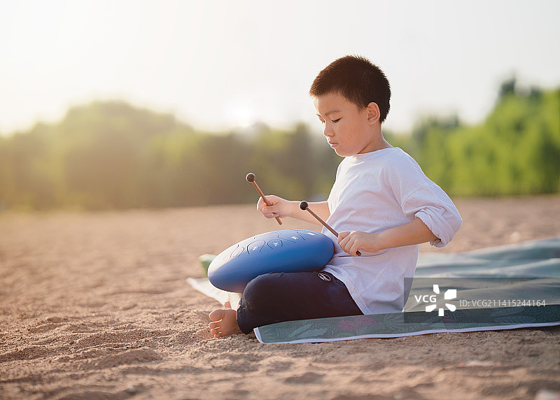 小男孩在夕阳西下的河边的沙滩上拿着鼓槌演奏空灵鼓图片素材