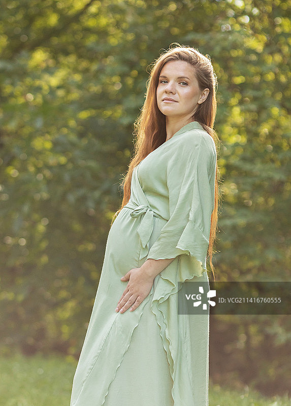 年轻孕妇站在公园里看着镜头，周围是绿树图片素材