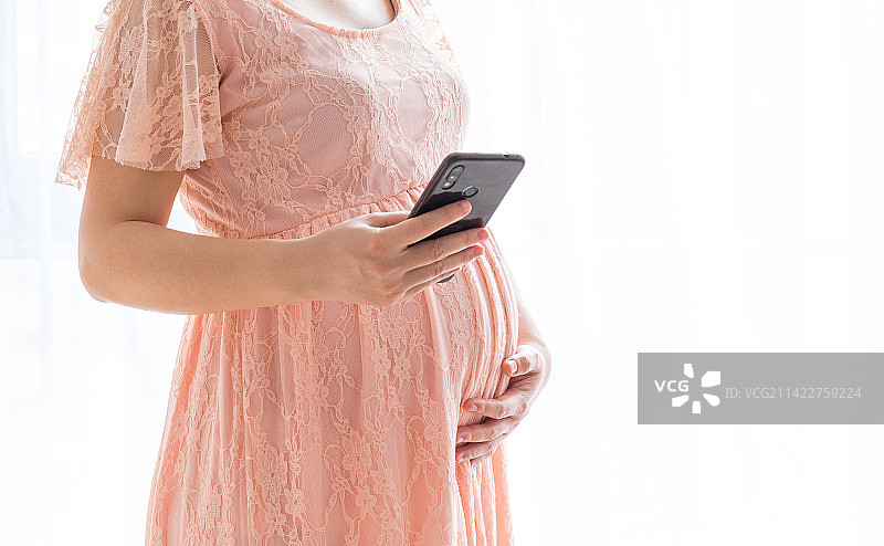 一名年轻孕妇在窗边玩手机图片素材