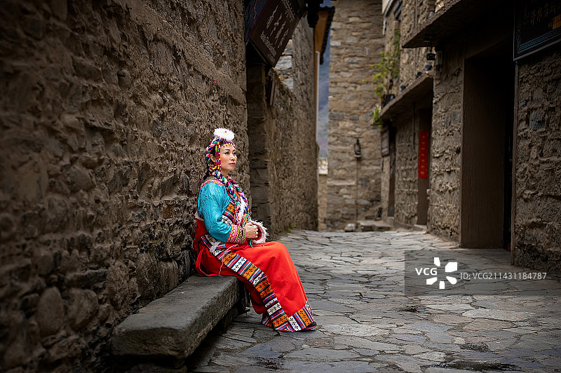 一位穿着藏族服装的妇女坐在四川阿坝州古老的甘堡藏寨的一条小巷的石头凳子上图片素材