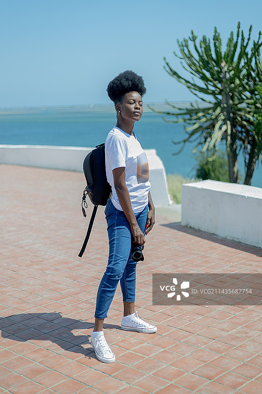在安哥拉罗安达，穿着休闲服装，背着背包，站在清澈蓝水和天空背景下的微笑年轻女人肖像图片素材