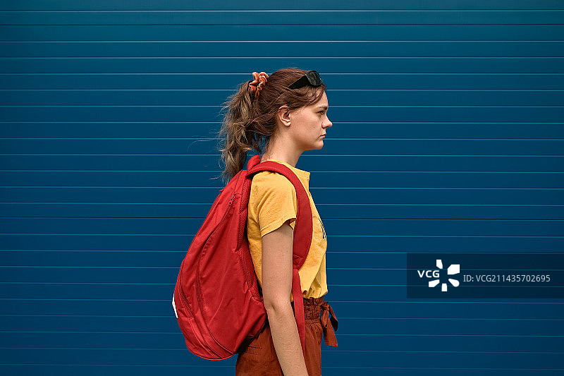 背红色背包的年轻女子靠在蓝色墙壁上的侧面照，俄罗斯图片素材