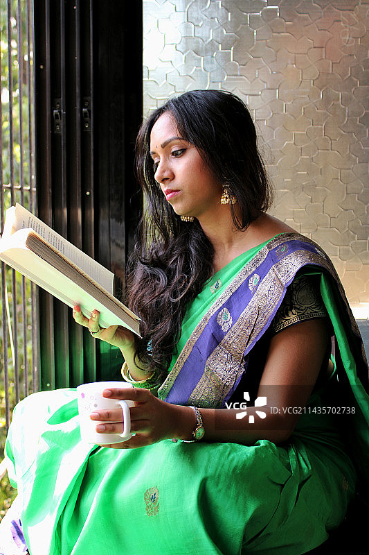 穿着绿色连衣裙靠窗阅读的微笑年轻女性肖像图片素材