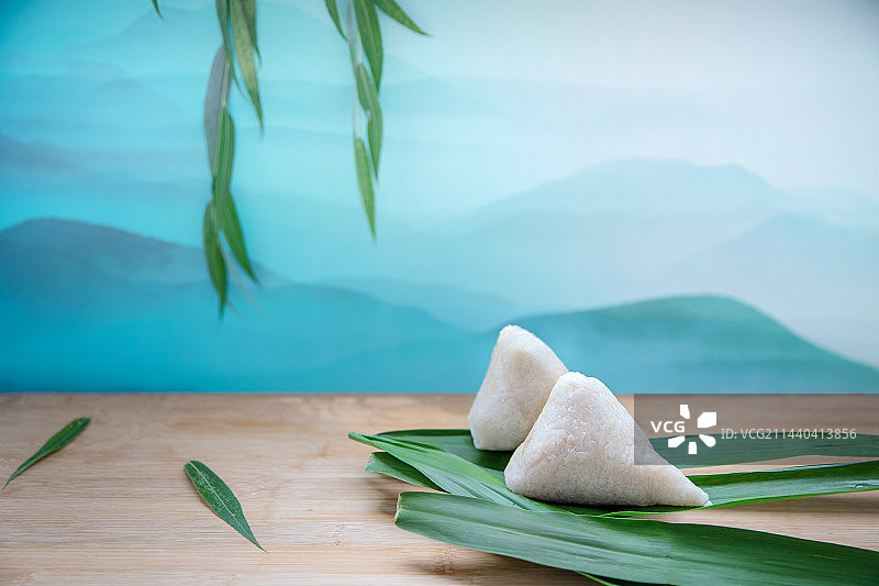 蓝色群山背景木纹桌面绿色粽叶上两只煮熟的三角形白米粽子图片素材