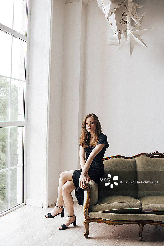 身穿黑色连衣裙的年轻女孩坐在俄罗斯窗前的沙发上图片素材