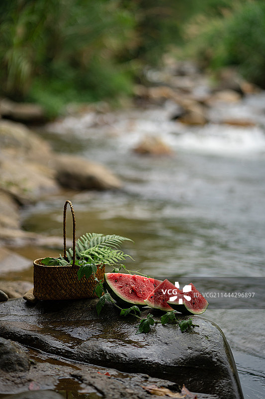 户外溪边石头上的消暑佳品西瓜与装满绿植的篮子图片素材