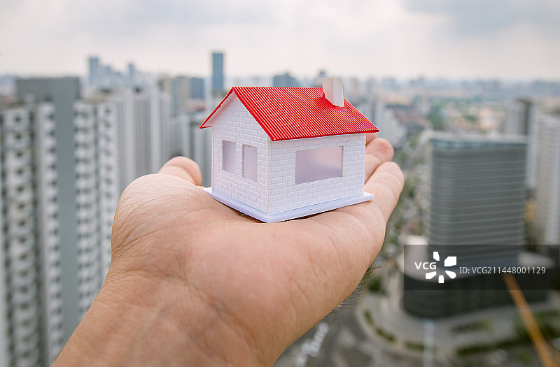 城市楼房背景下手托着一个房子模型：房地产 楼市政策概念图片图片素材