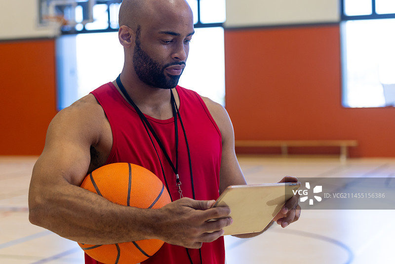 专注的混血男教练拿着篮球并在健身房使用平板电脑图片素材