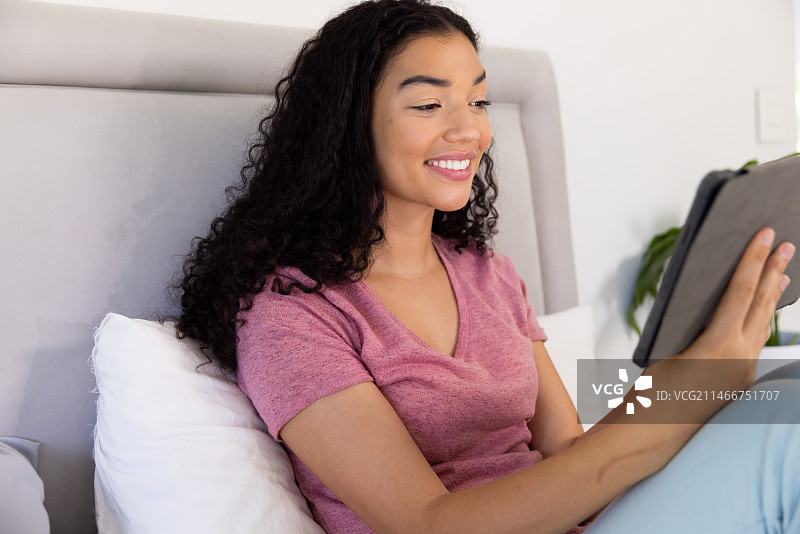 快乐的混血女人在阳光明媚的家里使用平板电脑坐在床上图片素材
