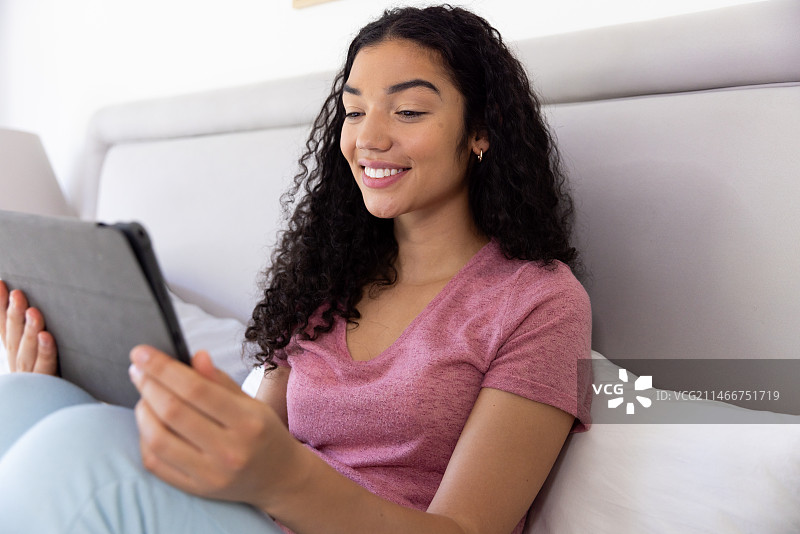 快乐的混血女人在阳光明媚的家里使用平板电脑坐在床上图片素材