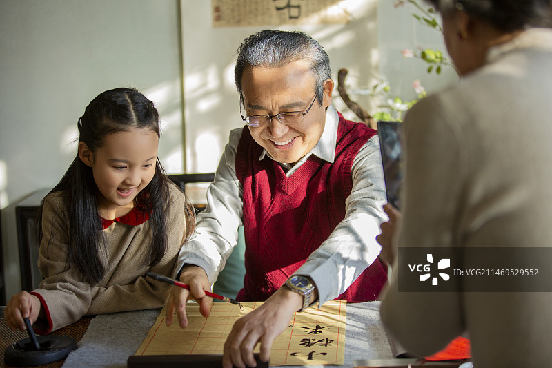 东方家庭过年：爷爷教孙女在书房写毛笔字，奶奶给他们拍照、录视频图片素材