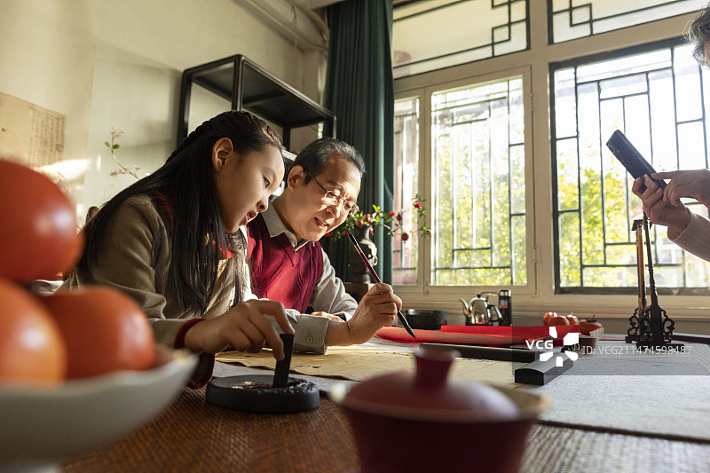 东方家庭过年：爷爷教孙女在书房写毛笔字，奶奶给他们拍照图片素材
