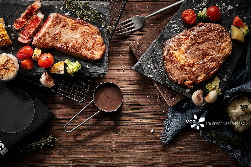 熟的牛排放在深色的木板上，旁边配有水果蔬菜和刀叉图片素材