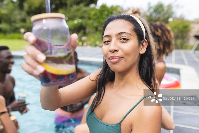 穿着绿色泳衣的年轻混血女人拿着水果饮料，在泳池派对上微笑图片素材