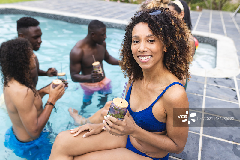 穿着蓝色泳装的混血女人在泳池边享用饮品；不同的朋友在背景中聊天图片素材