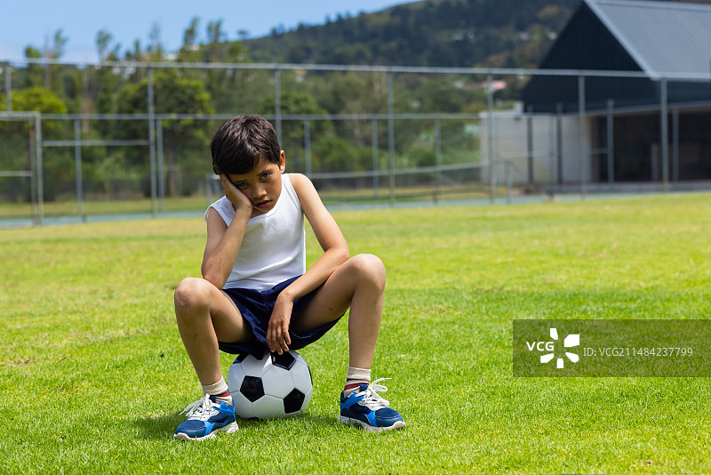 混血男孩坐在足球上，在阳光明媚的学校操场上看起来很悲伤，有文案空间图片素材