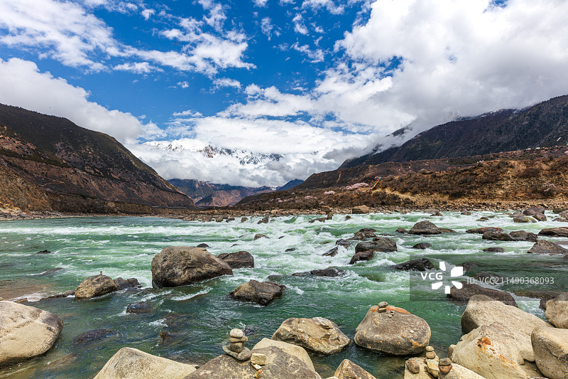 西藏林芝雅鲁藏布江大峡谷，绿色江水奔腾，山坡上桃花盛开，远处云雾笼罩南迦巴瓦峰雪山，岩石上有玛尼堆图片素材