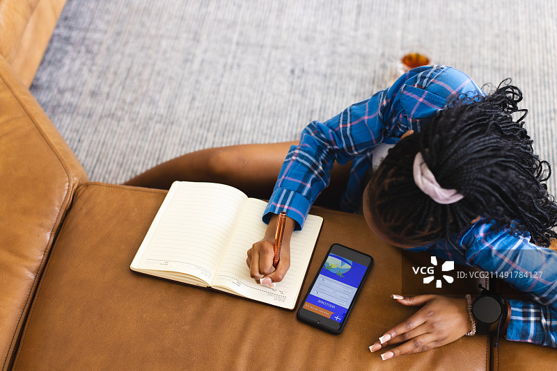 年轻的非裔美国妇女穿着蓝色格子衬衫在家里的笔记本上写作，有复制空间图片素材