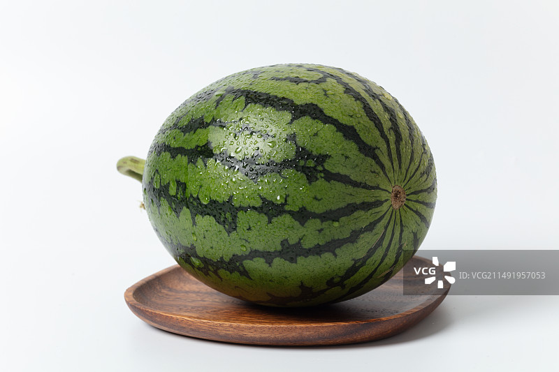 白色背景上，一个带水珠的绿皮西瓜平放在木盘里图片素材