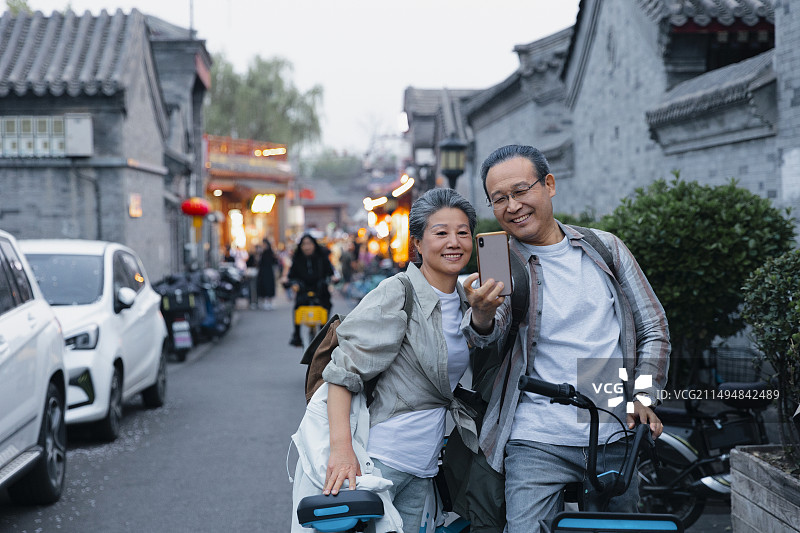 老年人City walk系列:老年夫妇在北京什刹海胡同骑着共享单车自拍图片素材