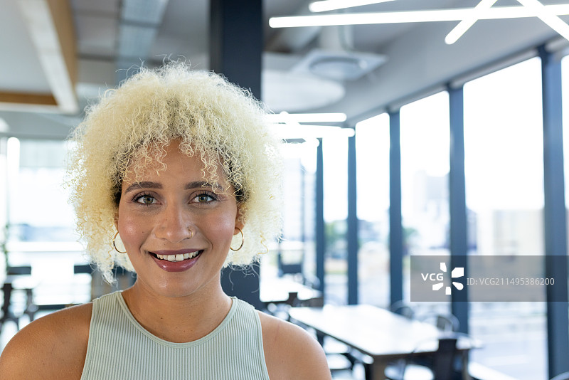 混血女人，金色卷发，笑容满面，在办公室里穿着浅色上衣和圈环耳环，有复制空间图片素材