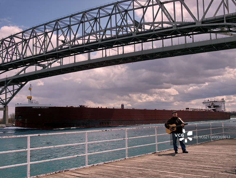 货轮在蓝水大桥下行驶，连接着密歇根州休伦港和安大略省萨尼亚，男子在木板路上弹吉他图片素材