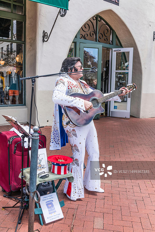 模仿猫王的人穿着猫王式的白色亮片服装，在人行道上手拿麦克风弹吉他唱歌图片素材