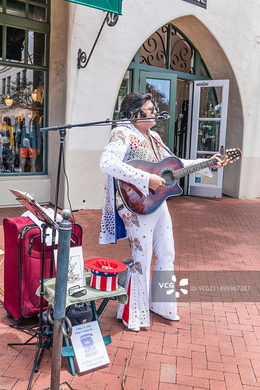 模仿猫王的人穿着猫王式的白色亮片服装，在人行道上手拿麦克风弹吉他唱歌图片素材