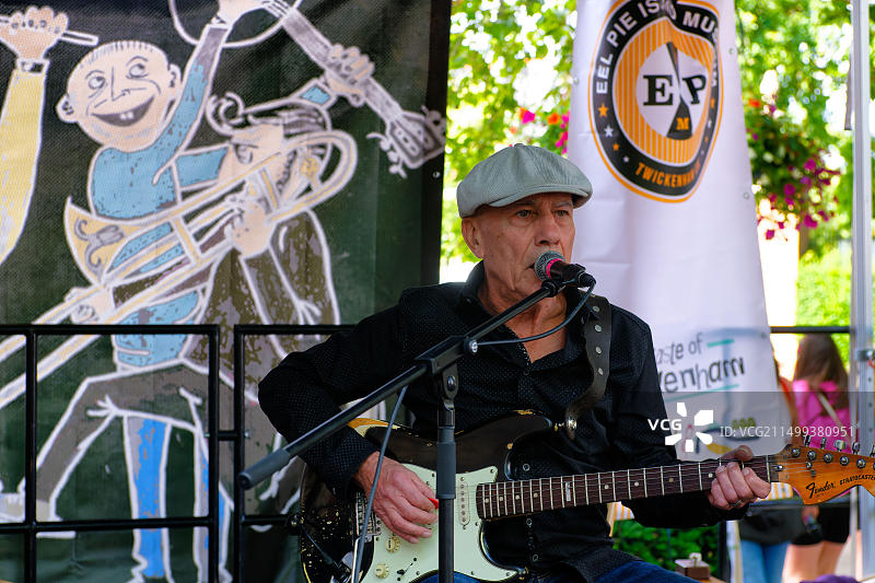 在特威克纳姆美食节上，吉他手和歌手坐在舞台上的麦克风前，戴着扁帽图片素材