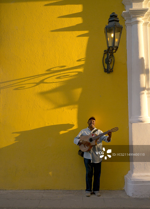 在古巴哈瓦那，一位西班牙裔男街头音乐家在下午晚些时候对着一面黄色墙壁弹吉他，有强烈的阴影图片素材