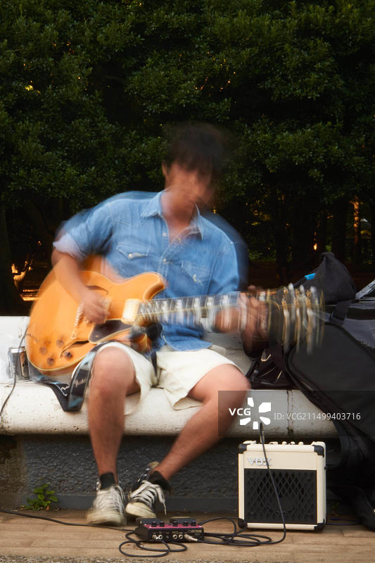 在东京涩谷代代木公园，一位穿着牛仔衬衫的二三十岁日本男人坐在长椅上弹奏黄色电吉他。图片素材