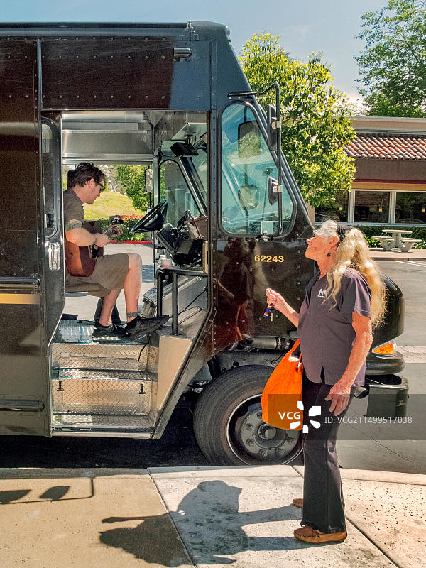 美国联合包裹运送服务公司（UPS）司机在加利福尼亚州拉古纳尼格尔的购物中心送货间隙弹吉他，一位路人欣赏他的音乐图片素材