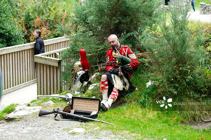 在格兰达洛山谷修道院遗址入口处，身着苏格兰士兵服装的男子坐在盔甲旁吹奏风笛图片素材