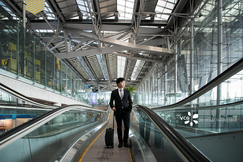 亚洲年轻商人拉着行李箱在机场乘坐自动扶梯。商务旅行概念。图片素材