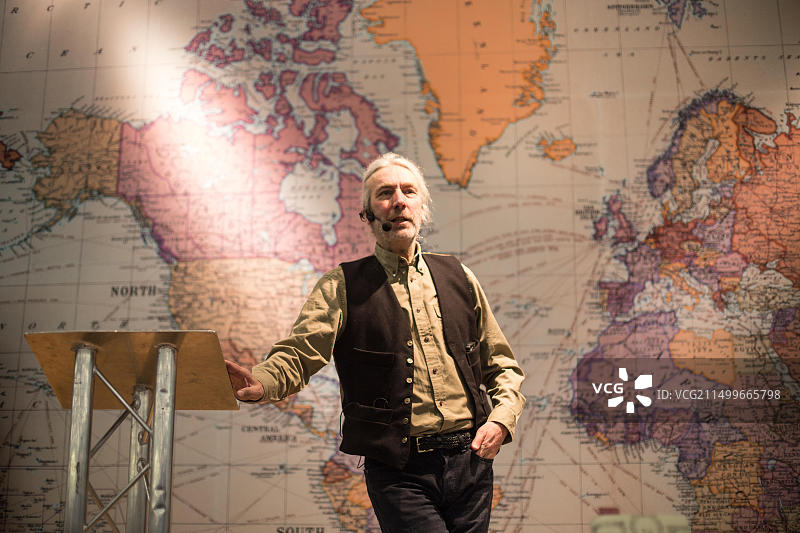 西蒙·巴恩斯在肯辛顿奥林匹亚目的地旅游展上演讲，背景是一幅巨大的世界地图图片素材