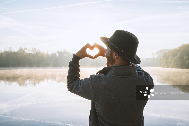 年轻男人在日出时站在湖边，用手指在美丽的风景上做成心形画框，水中倒影。人们热爱旅行图片素材