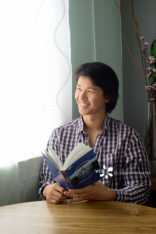 亚洲年轻男人坐在桌旁，手拿旅行书，面带微笑，望向窗外图片素材
