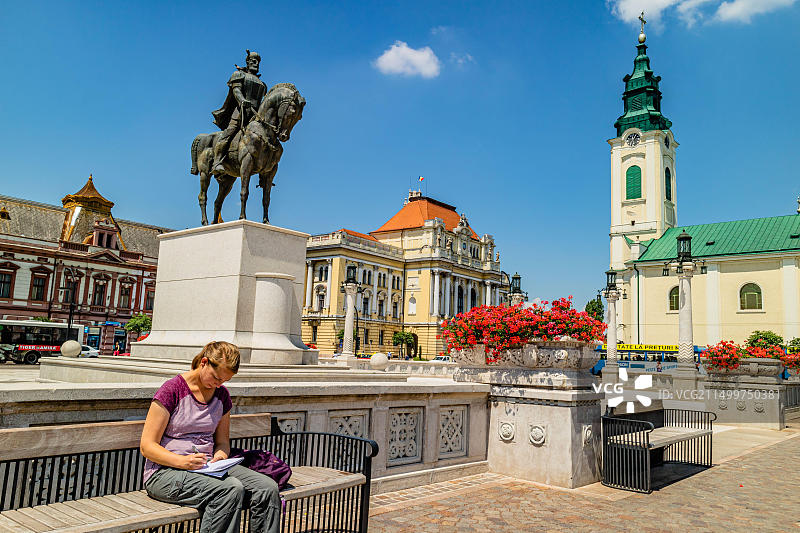 一位女士坐在长椅上写作，周围是巴洛克和分离派建筑。罗马尼亚奥拉迪亚市团结广场，2017年6月。图片素材