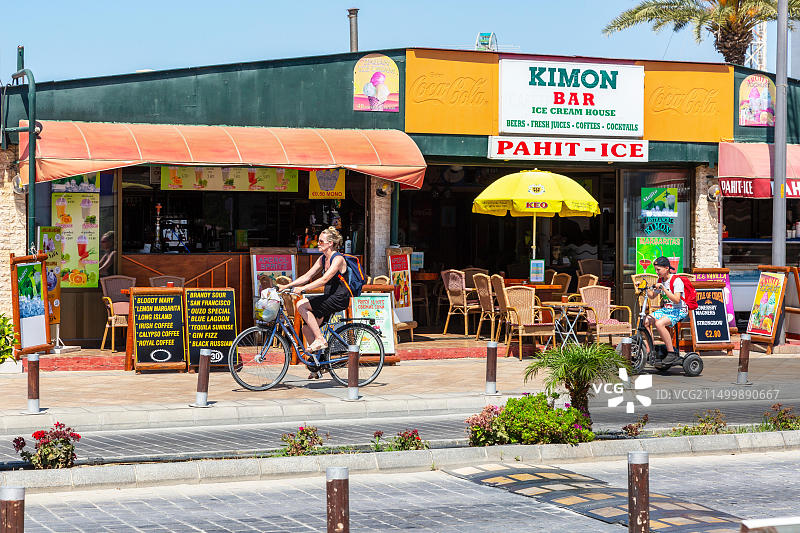 塞浦路斯阿依纳帕公共酒吧前，儿子骑着电动三轮车，女人骑自行车在自行车道上图片素材