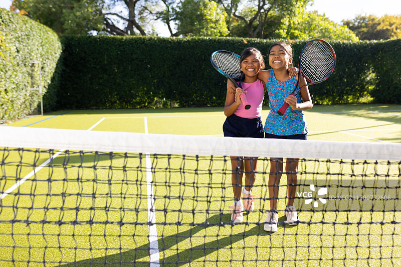 户外，两个混血小姐妹在网球场上笑着拿着网球拍图片素材