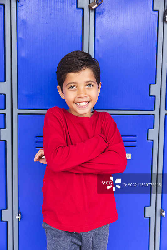校园里，年轻的混血男学生在户外倚靠着蓝色储物柜，双臂交叉图片素材