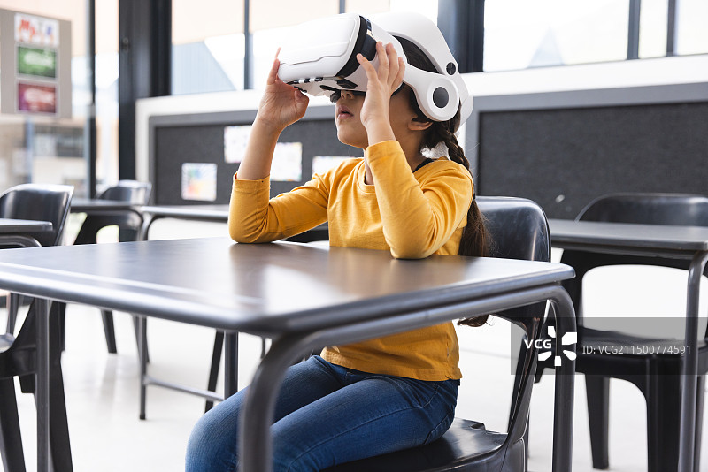 在学校里，年轻的混血女孩戴着VR头显坐在教室的桌子旁图片素材