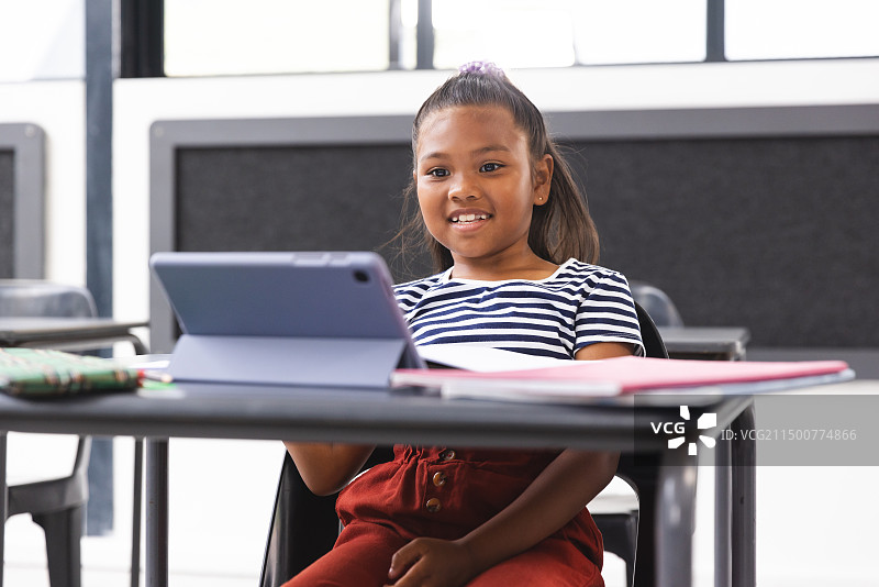 在学校教室里，年轻的混血女孩坐在桌子旁看平板电脑图片素材